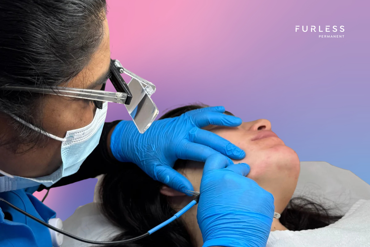 Electrologist in blue uniform wearing gloves and a mask performing electrolysis on a female client's jawline.