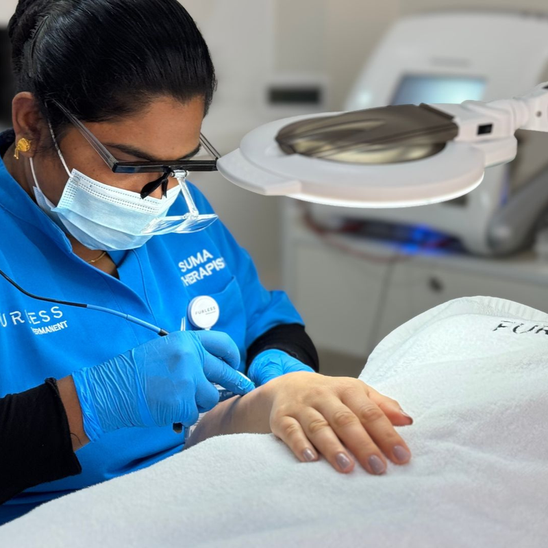 Electrologist in blue uniform wearing gloves and a mask, doing electrolysis on a patient's arm.